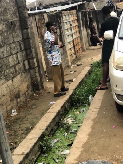 A street scene features a drainage ditch choked with green aquatic plants and litter. A person stands beside the ditch, clad in tan pants and a patterned shirt, using a phone. Nearby, another person in a black shirt stands by a white car. A third individual lies on a concrete step in the background. The surroundings include brick walls and buildings with metal grilles, indicating an urban environment.
