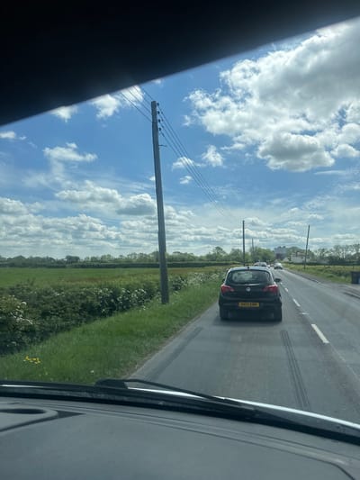 The image, taken from inside a vehicle, shows a view of a road in East Huntspill, United Kingdom. A dark-colored hatchback car is driving on the right side of the road. To the left of the road is a green field with some trees and power lines extending across the sky. The sky is blue with many white clouds. The vehicle from which the photo is taken is partially visible in the foreground.