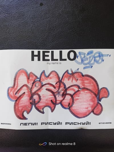 The image shows a close-up, eye-level view of a piece of paper with drawings and text on it, placed on a black, textured surface, possibly a leather-bound book or a car seat. The paper has a white background and features a bold, black text reading "HELLO" on the upper left side. To the right of "HELLO," there is a smaller, handwritten phrase in blue marker that appears to be a stylized signature or tag. Below "HELLO," in smaller black font, is the text "my name is."
Dominating the lower portion of the paper is a colorful graffiti-style drawing rendered in red and outlined in dark blue and black. The drawing is abstract, with flowing, bubble-like shapes and sharp, angular elements, suggesting a stylized word or character.
Along the bottom edge of the paper, there are several text elements. On the left, a small logo or text reads "BOMMAN". In the center, in black block letters, are the Russian words "ПЕПИ! РИСУЙ! РИСКУЙ!", which translate to "Pepi! Draw! Risk!". On the right, in smaller black letters, is the word "STICKERS".
The image also contains a watermark in the bottom right corner that reads "Shot on realme 8" along with a colorful infinity symbol. The overall impression is of a creative piece, possibly a sticker design, presented in a casual setting. There is no indication of the city of Shelekhov, Russia, within the image itself.