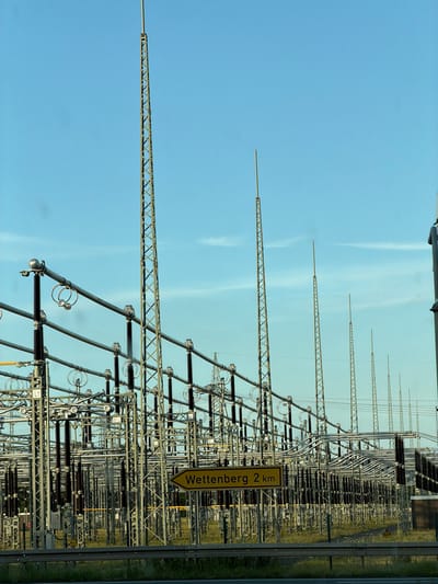 A yellow directional sign in the city of Gießen, Germany, points to "Wettenberg 2 km". The sign is situated in front of a large electrical substation complex, characterized by numerous metal towers, high-voltage power lines, and insulation equipment. The sky is clear and blue, with a few wispy clouds. The ground is covered in green grass and a wire fence encloses the substation. No people are visible in the image.
