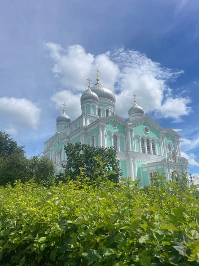 A wide-angle, low-angle shot captures a vibrant turquoise and white Orthodox church, partially obscured by lush green foliage in the foreground. The church features multiple silver domes topped with golden crosses, and its facade is adorned with arched windows and decorative architectural details. The sky above is a bright blue, dotted with fluffy white clouds. The church is identified as the Trinity Cathedral, located in the village of Diveyevo, Russia.
