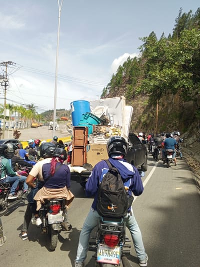 The image captures a traffic jam on a road in San Antonio de Los Altos, Venezuela, under a bright, partly cloudy sky. The scene is dominated by a long line of motorcycles, many carrying multiple passengers and significant amounts of cargo. One truck in the foreground is overloaded with various items, including a large blue barrel, what appears to be a mattress, and several boxes, one of which displays the text "Premium". The riders and passengers are wearing helmets, indicating a focus on safety. The road is paved and appears to be a two-lane highway. On the left side of the road, there are palm trees and some buildings, suggesting a semi-urban or peri-urban environment. The right side of the road features a steep, vegetated hillside with numerous trees. The time of day is likely daytime, given the strong sunlight and shadows. The overall mood is one of everyday commuting or perhaps people transporting goods, common in this region. One motorcycle license plate visible reads "LJ8C84R" and another visible plate contains the text "REPUBLICA BOLIVARIANA DE VENEZUELA AH9N49E BARINAS".