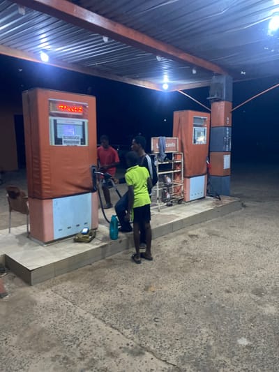 The scene depicts a nighttime view of a fuel station located in Sasaro, Nigeria. The area is illuminated by several bright, exposed light bulbs affixed to the underside of a corrugated metal roof structure, supported by orange metal beams. The ground surface is a coarse aggregate concrete.
In the foreground, there are two fuel dispensers. The left dispenser is orange on its upper section and light blue on its lower half. Its digital display shows red characters and a lower panel with "8229955438" and "HOMEPOINT" text. A fuel nozzle from this dispenser is actively in use. A yellow plastic jerrycan and a blue plastic bottle are placed on the ground near its base. A brown plastic chair is positioned to the left of this dispenser. Three males are present. One male, wearing a red t-shirt, stands behind the left dispenser. Another male, in a bright green t-shirt and dark shorts, stands in front of the left dispenser, facing right, with his hands near the engaged fuel nozzle. A third male, wearing a dark top with white stripes, stands to the right of the male in green, adjacent to a metal utility cart.
The right fuel dispenser is also orange and light blue, and its nozzle is holstered. A metal utility cart with shelves, holding several small containers (likely lubricants) and draped with a grey cloth, is positioned between the right dispenser and a dark grey and orange pillar. This pillar displays "NO SMOKING" in vertical white text on its dark grey section. The background beyond the illuminated canopy is dark, indicating night.