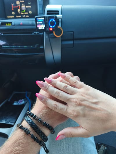 In an interior shot of a vehicle, likely in Tanowo, Poland, two individuals' hands are clasped together. The hand on the right, adorned with long, pointed acrylic nails featuring a French manicure with bright pink tips, belongs to a person with fair skin. The hand on the left, which is also fair-skinned and shows hair on the arm, is wearing three stacked bracelets: a braided black leather band in the middle, and two beaded bracelets on either side. These beaded bracelets consist of dark matte beads, interspersed with beads that appear to be wood or a similar material and small metallic cylinders.

The background reveals the dashboard of the vehicle. A car stereo system is visible, along with a digital display showing "9:38". Above the stereo, a small electronic device, possibly a dashcam or GPS unit, is mounted and displays various readings, including a blue circular light. The overall environment suggests a calm moment shared between two people inside a car.