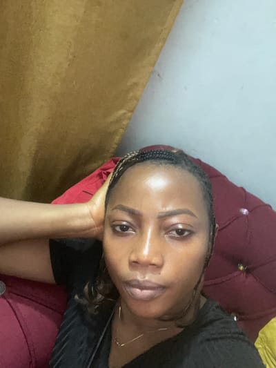 A close-up selfie shows a young woman with braided hair and a black t-shirt. She is resting her head on her left hand, which is propped on a red upholstered surface, possibly a couch or chair. Her right arm is bent and also resting on the red surface. The background features a golden curtain on the left and a light blue wall on the right. The lighting appears to be artificial, creating highlights on her forehead and cheekbones. The overall impression is a casual, indoor setting. The location in the city of Iyiowa Odekpa, Nigeria, is inferred from the context provided, but not visually evident in the image itself.