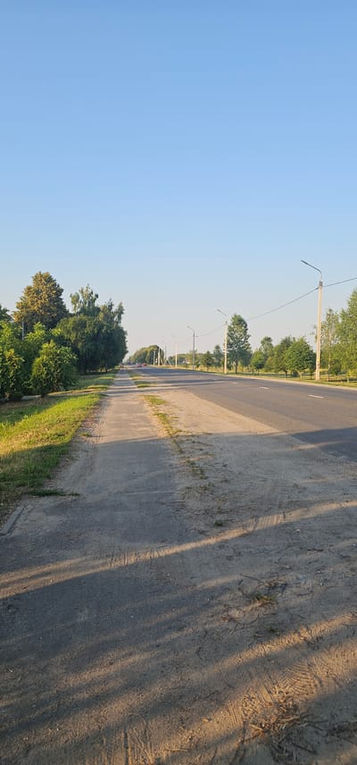 The image depicts a road lined with trees and streetlights in a rural setting. The road is paved and has white markings, suggesting it is a public thoroughfare. The right side of the road is bordered by a sandy shoulder with sparse vegetation. The left side features a grassy verge and a dense line of trees, which then transitions into a clear blue sky.
Several streetlights are visible along the left side of the road, spaced at intervals, indicating it might be a well-maintained route. The trees are lush and green, suggesting it is either spring or summer. The sunlight casts long shadows across the road and the sandy shoulder, indicating that the photo was taken during either sunrise or sunset, likely late afternoon or early morning. The sky is clear and bright, suggesting fair weather.
In the distance, there are a few vehicles on the road, and more trees can be seen. The overall scene conveys a sense of calm and tranquility, characteristic of a rural landscape. The location context provided, Буховіцкі сельскі Савет, Belarus, places this scene within a specific geographical area known for its natural landscapes. There is no visible text in the image. The image is oriented vertically.