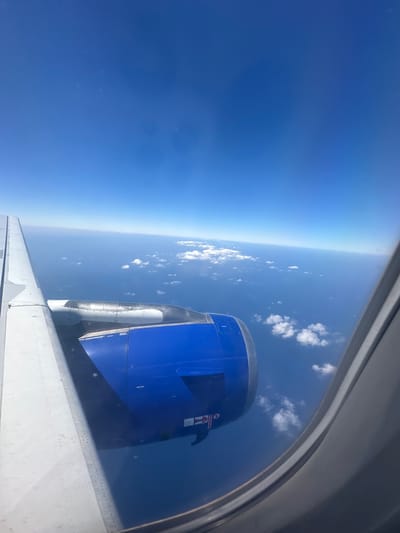 An aerial view shows the wing and engine of an airplane flying over a vast expanse of blue ocean dotted with scattered white clouds. The wing, partially visible on the left, is white and has visible rivets. The engine, predominantly blue, is situated below the wing. The horizon is a clear line separating the deep blue sky from the lighter blue ocean. A few fluffy clouds are visible in the distance, as well as some closer to the aircraft. The image is captured from inside the plane, with the edge of a window frame visible on the right. No specific location within Viehhausen, Austria can be determined from this image.