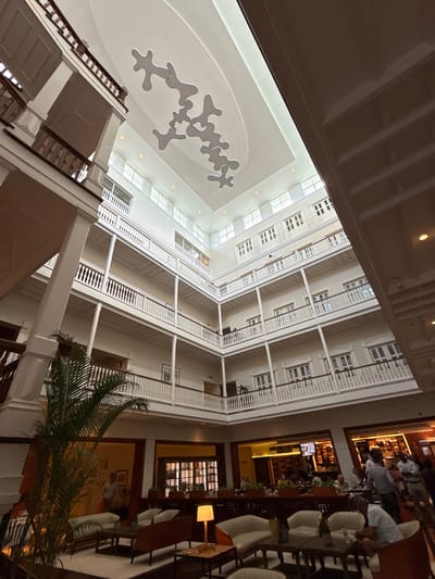 The image displays the multi-story interior atrium of the American Trade Hotel, located in Casco Viejo, Panama City, Panama. The scene reveals a central, open space surrounded by four visible floors of colonial-style balconies and corridors. Each level features white painted wooden balustrades with vertical slats and horizontal handrails, supported by white columns against light-colored walls. Numerous rectangular windows with multiple panes are visible on the upper floors. A large, rectangular skylight illuminates the atrium from above, featuring a suspended modern art installation composed of white, abstract, amoeba-like panels.
At ground level, a lounge area in the foreground and mid-left contains contemporary sofas and armchairs with light-colored upholstery and dark wood frames, arranged around low wooden coffee tables. A table lamp with an amber glow is present on one table. A large potted palm plant is positioned in the lower-left. To the mid-right, an elevated bar and restaurant area with dark wood finishes is visible. Approximately 10-15 individuals are present, standing or seated at the bar and tables, engaged in social interactions. Bottles and glassware are discernible behind the bar. The environment is illuminated by natural light from the skylight and warm artificial lighting from lamps and recessed fixtures.