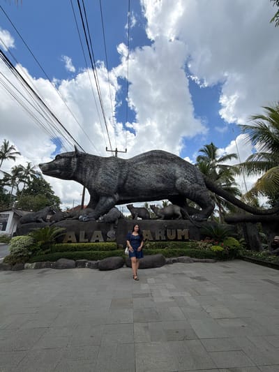 A woman stands to the right of a grey stone sculpture in Tegal Suci, Indonesia. The sculpture, featuring a large bear-like animal and other smaller figures, is positioned next to a sign bearing the text "ALAS RARUM." The area is paved with grey stone tiles and lined with greenery. Above, palm trees and electrical wires are visible against a backdrop of blue sky and white clouds.