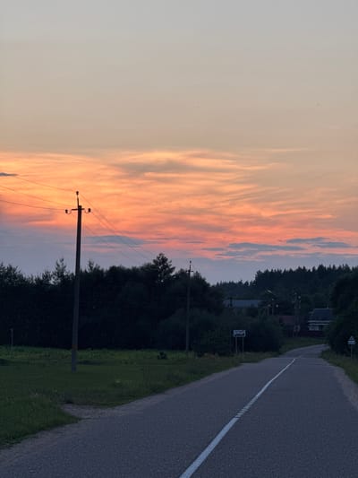 A sunset over a rural road in the Neman rural council, Belarus. The sky is filled with vibrant orange and pink clouds, transitioning to a pale blue at the top. A single utility pole with wires stands on the left side of the road. The road itself is asphalt with a white dashed line down the center. On the right side of the road, there are signs with Belarusian text, indicating a populated area. Trees line the background, and in the distance, houses with dark roofs are visible. The scene is serene and appears to be a quiet evening in the Belarusian countryside.