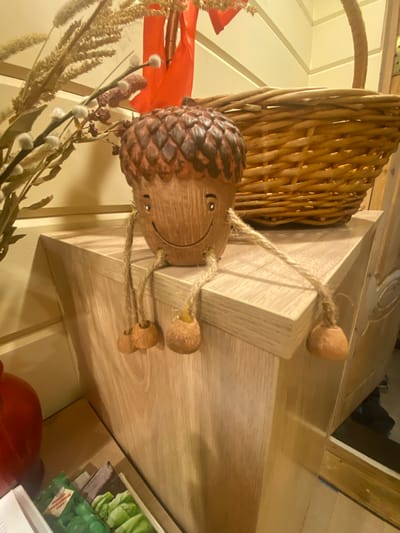 In Нарынка, Russia, an acorn figurine with a smiling face and dangling legs is perched on the edge of a wooden surface. The acorn is brown with a textured cap. Its legs are made of twisted rope, each ending in a small, round, acorn-shaped foot. Behind the figurine, a woven basket with a handle is visible. To the left of the acorn, a sprig of dried grass and pussy willows with white buds adds a touch of nature. A bright orange ribbon is also present in the background, suggesting a decorative or seasonal arrangement. The wooden surface on which the acorn sits appears to be part of a shelf or mantelpiece. Further to the left, a red, ribbed object, possibly a vase, is partially visible. At the bottom of the image, a stack of what appear to be seed packets with green imagery can be seen. The overall scene suggests an indoor decorative setting, likely a home or shop.