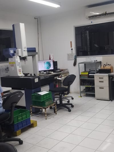 The image depicts an interior scene, likely a laboratory or workshop, in the city of Ciantra, Indonesia. A large, white and blue Coordinate Measuring Machine (CMM) dominates the left side of the frame. To the right of the CMM, a black office chair is positioned in front of a computer monitor displaying a circular graph. Behind the monitor, a metal shelving unit holds various items, including boxes and what appear to be tools or components. To the right of the shelving unit, a cream-colored filing cabinet with multiple drawers is visible. In the foreground, a green plastic crate with the text "PROGRESS: DELIVERY" is stacked on top of a blue plastic crate with text that is partially obscured but appears to say "PRDC RESG". The floor is tiled with white squares. The overall environment suggests an organized but functional workspace.