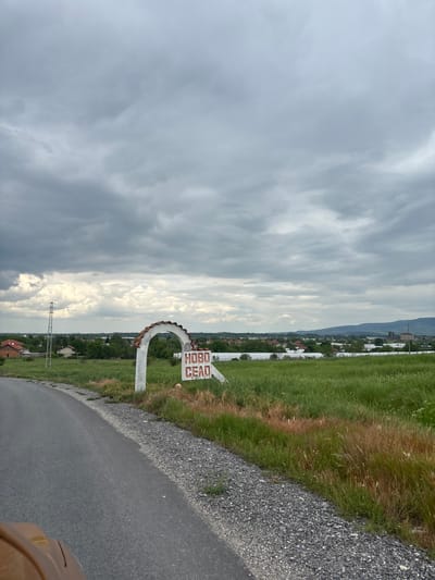 The image shows a road sign indicating the entrance to the village of Novo Selo (Ново Село in Cyrillic), located near Isperihovo, Bulgaria. The sign is white with red Cyrillic letters, and is situated at the edge of a paved road. Adjacent to the road is a patch of grassy verge and gravel. Beyond the road and sign, a rural landscape is visible, with fields, houses, and a distant mountain range under a cloudy sky. Power lines are visible in the midground. The photo is taken at an angle, suggesting it was taken from a moving vehicle.