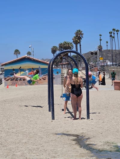 The image depicts a sunny day at San Clemente State Beach in California. In the foreground, a woman in a black swimsuit is swinging on a swing set. She has blonde hair and is wearing a turquoise helmet. Behind her, a young girl in a green shirt and shorts is also on a swing, her feet dangling as she moves. The swing set has a blue frame.

In the background, there is a mural painted on a wall that appears to depict surfers and waves. Palm trees are visible, and a bright blue sky suggests fair weather. There are also railway tracks with signals, indicating the proximity of a train line. Further in the distance, a hillside with sparse vegetation can be seen. The ground is covered in sand and gravel.

The overall atmosphere seems relaxed and recreational, fitting for a beach environment. The lighting suggests it is midday. Text is visible on the mural, spelling out "SAN CLEMENTE," and a yellow sign with "RAILROAD" and "TRAIN" is also present.