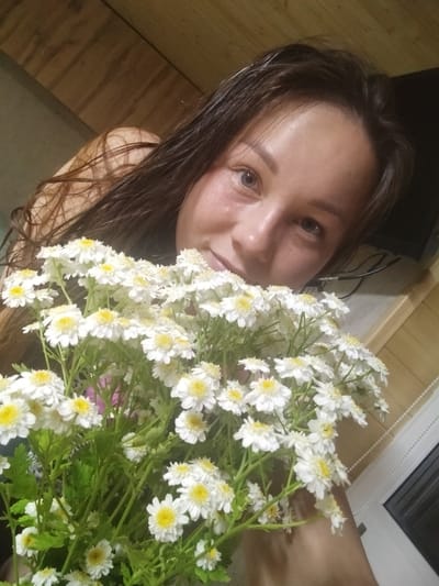 A young woman with long, dark, wet hair is holding a bouquet of white and yellow daisies. Her head is tilted, and she is looking towards the camera with a slight smile. The background appears to be an interior setting, with wooden paneling on the wall. There is a window with a white frame and a dark pane to the right of the woman. A dark object, possibly a television or monitor, is visible in the background behind her. The lighting is dim, creating a soft ambiance. The woman is wearing a dark garment, partially visible. The scene is a close-up, focusing on the woman and the flowers. The image was taken indoors. There is no visual information that indicates the location in the city of Вазерки, Russia.