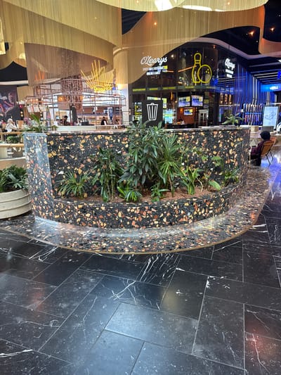 The image shows an interior view of O'Learys, a sports bar and restaurant located in Leidschendam, Netherlands. The foreground features a curved, dark-colored planter filled with various green plants, constructed from a material resembling terrazzo with colorful chips. The planter is set on a black marble-tiled floor with white veining. In the background, the O'Learys establishment is visible with its distinctive yellow bowling pin logo. The interior appears to be a lively entertainment venue, with bright lighting and multiple screens displaying content. A woman is seated in a brown chair to the right, facing away from the camera. Another person is seated at a table in the distance, and several other individuals are seen in the background, some of whom appear to be staff members. The overall environment suggests a modern and dynamic entertainment and dining space.