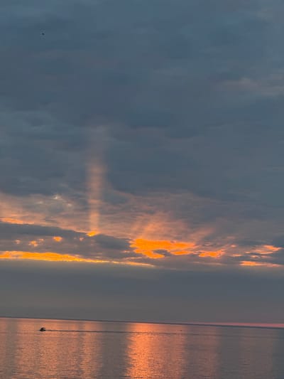 A wide shot captures a serene seascape at dusk near Ustka, Poland. The sky is a canvas of dramatic clouds, with streaks of vibrant orange light piercing through the gray masses. Sunbeams emanate from behind a cloud bank, creating an ethereal glow that illuminates the water below. A solitary boat is seen in the distance, leaving a subtle wake as it traverses the calm, reflective surface of the sea. The overall atmosphere is one of peaceful transition from day to night.