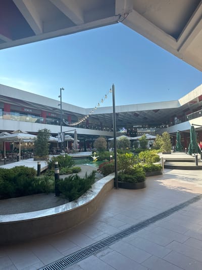 This image depicts an outdoor area within a modern shopping center. The scene is characterized by sleek architecture with concrete overhangs and multi-level buildings housing shops. A pathway, paved with light-colored tiles, runs along the right side of the frame, featuring a long, narrow metal drain. Lush greenery is incorporated into the design, with well-maintained bushes and small trees planted in curved planters. One planter prominently displays a bright yellow duck sculpture in a small water feature.

The architecture suggests an open-air mall or a similar commercial complex. The presence of tables and chairs outside some establishments indicates that dining or cafe areas are available. Though the image is vertically oriented, it appears to be taken during the day, with clear blue skies visible and shadows cast by the structures, suggesting sunny weather. The overall atmosphere is peaceful and orderly, conveying a sense of a pleasant public space.

Visible text includes "SPORTİF" and "DEMLİK" on storefronts, indicating specific businesses within the center. The location context is Çanakkale Merkez, Turkey. The image captures a moment of quietude, with few people visible in the distance, suggesting it might be early in the day or a less busy period.