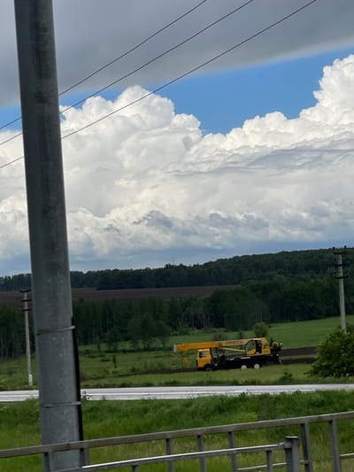 The image shows a scene in Кокино, Russia. A yellow mobile crane is parked on a road, alongside a green field and a treeline. Above, there are cumulus clouds filling the sky, with a blue patch of sky visible. In the foreground, a metal structure partially obscures the view. Power lines also cross the sky.