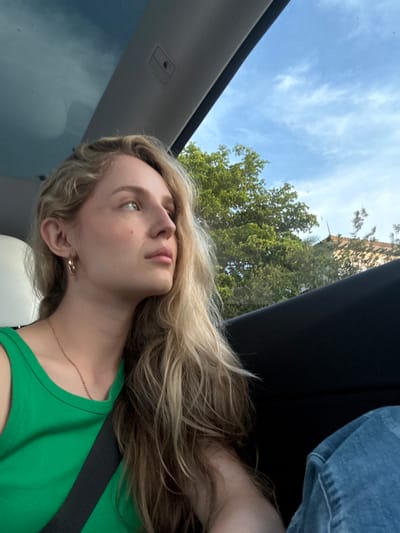 A blonde woman with light skin and a fair complexion is seated inside a vehicle, looking to her left. She is wearing a green tank top and a gold necklace. Her hair is long and wavy, falling over her shoulder. A black seatbelt is visible across her chest. The interior of the vehicle appears to be modern, with a panoramic sunroof and a dark interior panel. Outside the vehicle, lush green trees and a building with a terracotta roof are visible against a bright blue sky with scattered clouds. The scene suggests a sunny day in a suburban or urban environment. The image was taken in Coral Gables, United States of America.