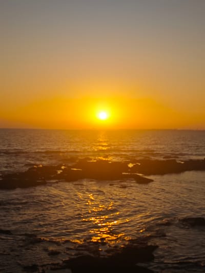 The image depicts a sunset over the ocean at Costa Oeste, Spain. The sun, a bright orb of yellow and orange, is low on the horizon, casting a warm glow across the sky and water. The sky transitions from a pale grey at the top to a vibrant orange closer to the sun. The ocean surface is calm, with small waves gently lapping against dark, rocky shorelines that are partially submerged in the water. The sun's reflection shimmers on the water, creating a path of golden light. The scene evokes a sense of tranquility and natural beauty. There are no people, objects, or text visible in the image. The time of day is clearly sunset, and the weather appears clear. The location cues from the prompt specify Costa Oeste, Spain.