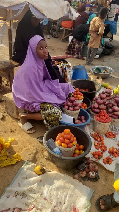 The image depicts a market scene in Kawo, Nigeria. A young woman wearing a lilac headscarf and robe sits near buckets filled with produce for sale. The buckets contain red onions and red tomatoes. Other produce, including yellow tomatoes and red peppers, is spread out on a sheet on the ground nearby. The ground is sandy and there are other people and structures visible in the background.