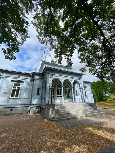 The image depicts a light blue wooden house with a white porch and a grand staircase leading up to its entrance. The house has several windows and ornate architectural details. The surrounding area is covered with gravel and fallen leaves, suggesting an autumn setting. Lush green trees frame the scene, with their branches partially obscuring the sky, which is partly cloudy. In the background, another building with a yellow facade is visible. The house appears to be a historic structure, possibly a museum or a cultural landmark. This scene is likely the Vyborg-based artist Alexander Volkov's house-museum in Sortavala, Russia, known for its distinctive architecture and connection to the artist's life and work.