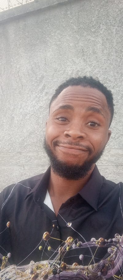 The image displays a male individual, appearing to be in his 20s or 30s, positioned against a light grey, textured stucco wall in Tunga Mekidi, Nigeria. He has dark skin, short black hair, and a full black beard. His facial expression is a closed-mouth smile, with his eyes looking directly at the viewer and slight horizontal lines visible on his forehead. He is wearing a dark-colored collared shirt, likely black or dark blue, with a white undershirt visible at the collar. The dark shirt features an intricate abstract pattern of interconnected spheres and lines in various metallic and muted tones (gold, silver, purple, green). The background wall has a consistent texture, and a faint, blurred horizontal line is visible across the top portion of the image, suggesting a structural element above. The scene is evenly illuminated, indicative of daylight.