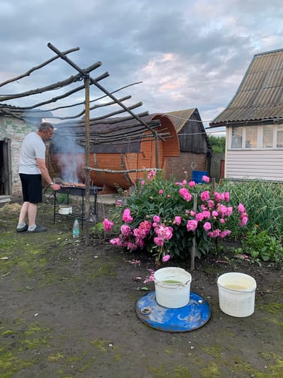 In Блеўчыцкі сельскі Савет, Belarus, a man is grilling meat on a portable barbecue. He is wearing a white t-shirt and black shorts. The barbecue is located on a muddy, overgrown yard. In the background, there are several wooden structures, including a barrel-shaped sauna and a small house with a corrugated roof. A bush with pink peonies is in bloom next to the barbecue. Two white buckets filled with water are on a blue metal barrel in the foreground. The sky is cloudy with hints of pink and orange from the setting sun.