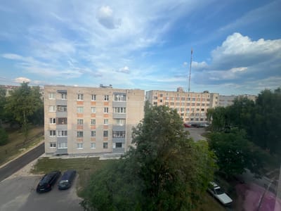 The image is an outdoor, eye-level view of an urban environment in Kobryn, Belarus, on a day with a blue sky filled with scattered white clouds. The foreground is dominated by the lush green foliage of several large trees, some of which obscure parts of the scene.

In the middle ground, two multi-story apartment buildings are visible. The building on the left is constructed of light-colored brick with multiple rows of windows, balconies, and some visible air conditioning units. The building on the right is taller and also made of brick, with a similar arrangement of windows and balconies. A tall communication tower with an antenna at its apex stands in the distance, behind and to the right of the second apartment building.

Below the apartment buildings, a paved area appears to be a street or parking lot. Two dark-colored vehicles are parked side-by-side on the paved surface. Further to the right, a white car is parked on a grassy area adjacent to the paved surface. Some areas of green grass and patchy dirt are visible between the buildings and the paved areas. The overall impression is of a residential neighborhood within the city.