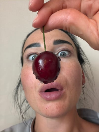 In this close-up, upside-down shot, a person's face and hand are the central focus, with a single, glistening cherry held between their fingers. The person has striking blue eyes, dark eyebrows, and light skin with freckles across their cheeks and nose. Their lips are slightly parted, revealing a hint of their tongue, and they appear to be wearing subtle makeup. A thin stem from the cherry extends towards the left side of the frame. The background is a neutral, light-colored surface, suggesting an indoor setting. The lighting is soft, highlighting the texture of the skin and the dewiness of the cherry. The image evokes a sense of playful intimacy and a focus on sensory experience, drawing attention to the natural beauty of the fruit and the person's features. The overall mood is one of casual sensuality and a moment captured in time.