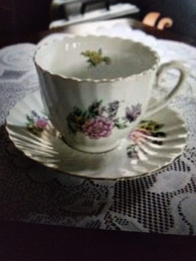 The image shows a white teacup and saucer with floral patterns, placed on a lace tablecloth. In the background, a dark surface with a lighter rectangular object is visible, possibly a piano or a table with some items on it. The lighting is dim, and the image is slightly out of focus. The scene does not provide any information about a specific location in Kualaraja, Indonesia.