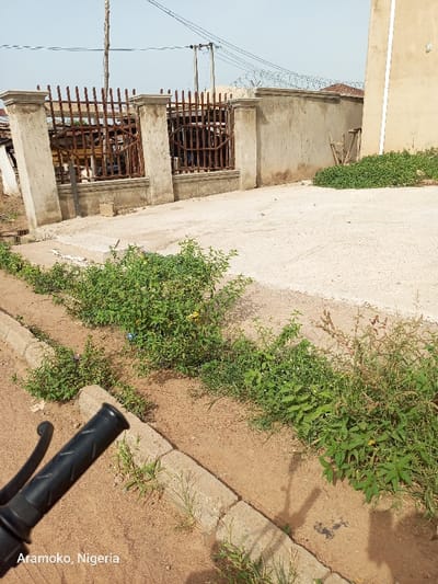 The image shows a dirt road with a concrete curb on the left side. The road is edged by a patch of green vegetation. On the right side is a concrete driveway, which leads to a concrete wall with a wrought iron gate. There is a building with a partial view of its side wall to the right of the driveway. The scene is a residential area in Ado Ekiti, Nigeria, near the coordinates 7.658° N, 5.233° E. The photo is taken from the perspective of a person standing near the concrete curb, with the handlebar of a motorcycle visible in the lower left corner. The scene is sunny, with the shadows cast by the vegetation and the building.