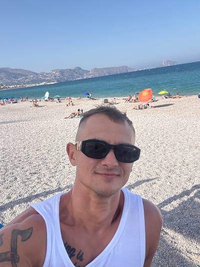 In Pla Parcial Platja, Spain, a man in a white tank top and sunglasses takes a selfie on a pebble beach. The turquoise ocean stretches behind him, meeting a hazy coastline dotted with buildings and mountains. Other beachgoers are scattered across the sand, some under umbrellas, while others lounge in the sun. The clear blue sky dominates the upper portion of the image.