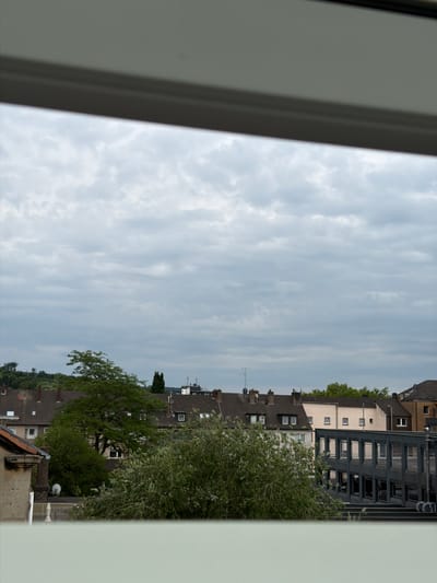This image, taken from an elevated perspective within the city of Witten, Germany, captures an overcast sky with scattered clouds. Below the sky, a panoramic view of residential and commercial buildings unfolds. The foreground shows the edge of an interior structure, possibly a window frame or sill.
Dominating the middle ground are several rows of terraced houses with dark-tiled roofs and visible windows. Green trees, some with dense foliage and others more sparse, are interspersed among the buildings, with one particularly large tree with a broad canopy prominently positioned. To the right, a modern, multi-story building with a dark facade and numerous windows is visible, featuring a distinctive architectural design with angled sections. The background reveals more distant buildings and a wooded hillside. The overall scene depicts a typical urban or suburban landscape under a cloudy sky. No people or specific actions are discernible in the image.