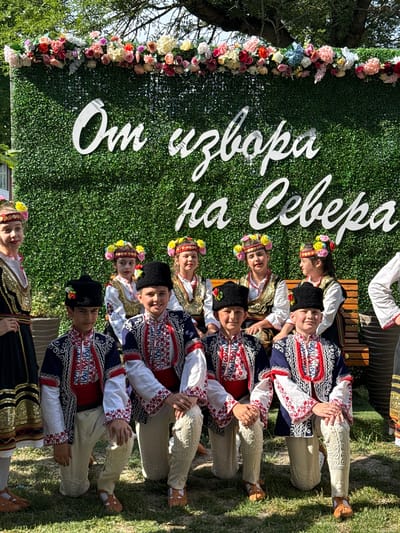 The image shows a group of children in traditional Bulgarian folk costumes posing in front of a green hedge wall adorned with white text and a floral garland. The text on the wall reads "От извора на Севера," which translates to "From the Source of the North." The children are dressed in brightly colored embroidered vests, shirts, pants, and hats, and some are wearing floral headpieces. The group appears to be participating in a cultural event or performance, likely held outdoors given the presence of trees and grass in the background. The event is taking place in the city of Pordim, Bulgaria, as indicated by the location tag.