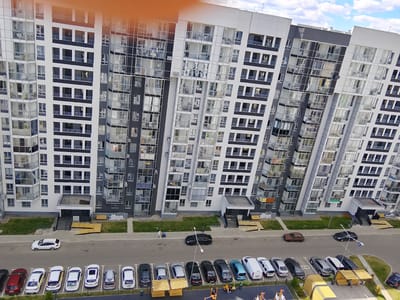 The image depicts a modern, multi-story residential building in Zhilino-1, Russia. The building features a contemporary design with a facade composed of white, gray, and dark gray elements, accented by numerous large windows and balconies. The architecture is characterized by sharp angles and a dynamic, asymmetrical composition.
In the foreground, a paved area serves as a parking lot and walkway. Several cars are parked, including a white SUV, a red sedan, a white sedan, a yellow sedan, and a dark-colored sedan. Further down the parking lot, more vehicles are visible, including a light-colored van, a white sedan, a silver sedan, a dark gray sedan, and a dark blue sedan. Two yellow wooden shelters with benches are present, with a few people seated beneath one. A dark-colored sedan is parked on the roadway, and a brown sedan is also visible.
To the left of the building, a grassy area with some landscaping is present. Two individuals are walking on the sidewalk, one in white clothing and another in darker attire. A second individual appears to be bending down near the first.
The sky above is partly cloudy, with white clouds scattered across a blue sky. The overall scene conveys a sense of urban residential living with a focus on modern construction.