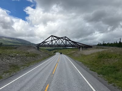 The image shows a view from a vehicle traveling on a two-lane asphalt road. The road has white solid lines on the outer edges and a dashed yellow line in the center. In the distance, a large truss bridge spans over the road. The bridge is dark in color and appears to be made of metal. Beyond the bridge, a hilly landscape with green grass, scattered rocks, and trees can be seen. The sky is partially cloudy with patches of blue sky visible. A few vehicles are visible on the road ahead, approaching the bridge. The overall environment suggests a rural or semi-rural setting. The location is identified as the E6 highway passing under a railway bridge near Trofors, Norway.