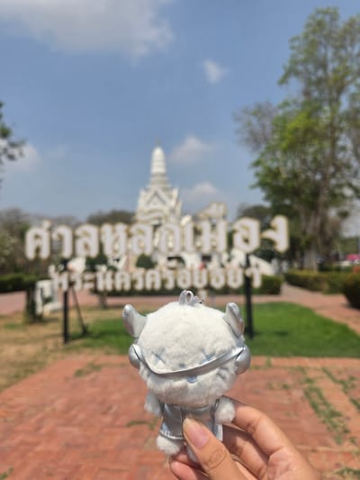 A hand holds a small, white stuffed animal against a background that features a large sign with Thai script and a white pagoda-like structure. The location is the Historic City of Ayutthaya, Thailand. The background also includes trees, grass, a clear blue sky, and brick walkways.