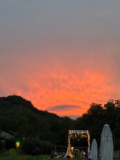 This is a landscape photograph taken outdoors at dusk, with the sky painted in vibrant shades of orange and pink from the setting sun. The scene is set against a backdrop of lush, dark green trees that form a hilly terrain. In the foreground and midground, there are signs of human activity and gathering.
Visible elements include several large, light-colored umbrellas, suggesting an outdoor event or seating area. A decorated archway, illuminated with strings of warm lights, is set up, implying a celebration or event. Small groups of people are discernible in the midground, some appearing to be interacting.
The time of day is clearly evening, as indicated by the dramatic sunset colors and the presence of artificial lighting. The weather appears calm, with the sky showing scattered, wispy clouds. The location context provided is Asureti, Georgia. The overall emotion evoked by the image is one of warmth, celebration, and the beauty of nature at twilight. No visible text is present in the image.