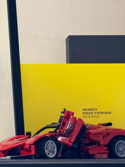 The image features a display with a yellow backing that reads "Museo Enzo Ferrari Modena" in black lettering. To the right, a red LEGO model of a Ferrari is partially visible, showcasing its intricate details, including wheels and engine components. The display is framed by a black border. The surrounding environment includes a white wall and a dark gray element, possibly part of a larger display structure. The location in Kafr Yasif, Israel, cannot be verified from this image alone.