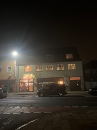 This nighttime image depicts a street scene in Bamberg, Germany, enveloped in a dense fog or mist that softens the light and obscures distant details. On the left, a multi-story building stands prominent. Its upper-floor windows are illuminated from within and adorned with decorative string lights, adding a festive glow to the hazy night.
The ground floor of the building hosts two commercial establishments. To the left, a brightly lit "PIZZA" restaurant displays a large sign featuring the word "PIZZA," a graphic of a character, and text indicating "Lieferservice" (delivery service) with partially visible phone numbers (e.g., "0951-..."). Adjacent to it is a business identified by "METZGEREI OLMA" (butcher shop), indicated by a sign with a star symbol. The text "METZGEREI OLMA" is also clearly visible on the side of a dark-colored van or car parked directly in front of this shop.
A strong, glaring street lamp is situated in the lower-left foreground, its light rays diffused by the fog. The street itself appears wet and reflective, stretching along the right side of the building, with a distinct paved sidewalk featuring a red-and-white curb marking. A bare, leafless tree stands between the building and the street, suggesting winter or late autumn. Parts of another vehicle are visible in the lower right corner. The overall scene is quiet and atmospheric, characterized by the interplay of artificial lights and the pervasive fog.