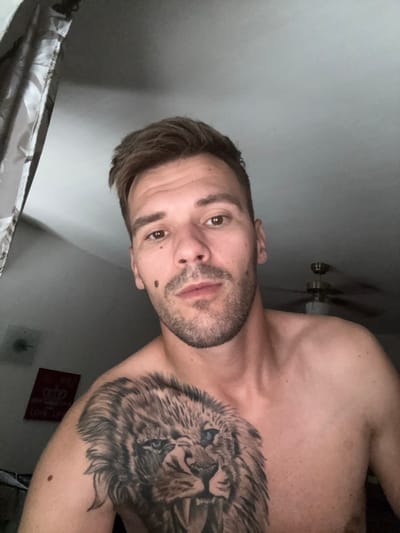 The media file is a selfie taken indoors, with the subject oriented sideways. The subject is a man with short, brown hair, a beard, and brown eyes. He has a mole on his left cheek and another on his upper lip. A prominent tattoo of a lion's head is visible on his chest and upper arm. He is shirtless, revealing his chest and shoulder.
In the background, a ceiling fan is visible, suggesting the room is indoors and likely in a warm climate or season. A patterned curtain is visible in the lower left corner of the image. A small, red square object with some text is affixed to the wall in the background, partially obscured. The text visible on this object appears to be "LOVE LIFE".
The lighting suggests the photo was taken indoors, possibly in the evening or with artificial lighting, as the ambient light is low. The overall mood appears casual and relaxed. There are no strong indicators of time of day beyond the indoor setting and artificial lighting. The location context provided is Sesto Calende, Italy, which is not directly discernible from the visual cues in the image itself, but serves as a contextual reference.