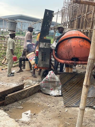 In Okinano, Nigeria, a construction site is captured under an overcast sky. Four individuals are present, engaged in activities related to a cement mixer. One man, wearing a light-colored outfit and red gloves, carries a large bucket of what appears to be cement on his head. Another man, in a light brown shirt and a cap, stands near the mixer. A third man, in a blue shirt, is seen interacting with the large orange cement mixer. The fourth person, wearing a sleeveless shirt and dark pants, sits on a white bucket holding another bucket. The cement mixer is positioned on a wheeled platform. The ground is muddy and uneven, with scattered debris, wooden planks, and a puddle of water. In the background, bamboo scaffolding surrounds a partially constructed building, and a modern building with glass windows is visible in the distance. The scene conveys a sense of ongoing construction work in a developing area.