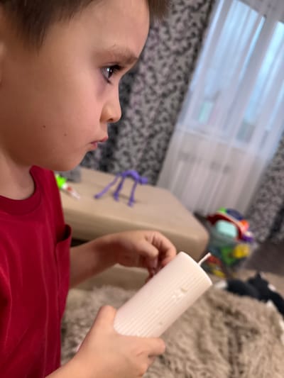 A boy is holding a white candle and appears to be looking at it intently. He is wearing a red shirt and is positioned in the foreground of the image. The background is blurred but appears to be a room with a window and some toys. The specific location in Malmyzh, Russia cannot be determined from the image.