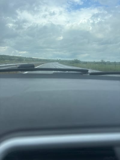 The image depicts a view from inside a moving vehicle, likely a car, on a road in Rogozinovo, Bulgaria. The road is wet, suggesting recent rain. The sky is overcast with clouds. The surrounding landscape consists of grassy fields. A portion of the car's interior is visible on the right side of the frame.