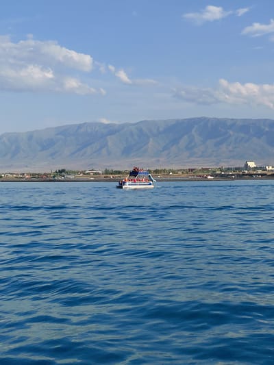 This media file captures a scenic view from a boat on a body of water, likely a lake, with a coastline and mountains in the background. The image is oriented vertically, suggesting it might be a rotated photograph.
On the boat, several people are visible, appearing to be enjoying a recreational activity. They are dressed casually and seem to be engaged in conversation or observing the surroundings. The boat itself is white with blue accents and has a railing. Life vests, in orange and red, are distributed among the passengers, indicating safety precautions.
The coastline features a line of buildings, suggesting a populated area, possibly a town or resort. Trees are scattered along the shore, and behind them, imposing mountains rise, their slopes appearing dry and somewhat arid. The sky is a bright blue with scattered white clouds, implying fair weather. The water is a deep blue with visible ripples, suggesting a gentle breeze.
The overall scene conveys a sense of leisure and a connection with nature. The bright sunlight and clear skies suggest it is daytime, likely late morning or early afternoon. The presence of mountains and a large body of water in a dry climate points towards a location like Akshi, Kazakhstan, as suggested by the location context. There is no visible text in the image.