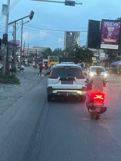 The image depicts a street scene in Kutaluhoksukon, Indonesia, characterized by moderate traffic and a somewhat unkempt environment. In the foreground, a white SUV with license plate BL 1607 EC is visible, followed by a motorcycle with a rider wearing a light brown shirt and pink pants, its taillight illuminated. The motorcycle's license plate is BL 4199 KAT. On the right side of the road, a billboard advertises "VEEVA GRAPE" for Rp 35,000. Other vehicles, including a white sedan with its headlights on and several motorcycles, are present in the middle ground. Several pedestrians are also scattered along the roadside. The road surface appears to be dirt or gravel, with debris and trash visible. Overhead, power lines and traffic signals are strung across the sky. The sky is overcast with a bluish-gray hue, suggesting it might be late afternoon or early evening. Buildings and trees line the street, with some appearing to be commercial establishments, including a sign for "Al...art." The overall atmosphere suggests a typical day in a bustling Indonesian town.