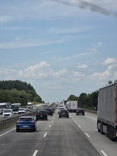The image depicts a multi-lane highway in Oberselters, Germany, with moderate to heavy traffic. The road surface is asphalt, marked with white lane lines. The weather appears to be overcast with scattered clouds, and a patch of darker clouds is visible in the upper portion of the image.
On the highway, there are numerous vehicles including passenger cars, vans, and trucks. In the foreground, a blue hatchback is in the right lane, and a large semi-trailer truck is in the adjacent lane to its right. Further ahead, traffic is denser, with vehicles in all visible lanes. Several cars and trucks are distinguishable, some with visible license plates.
To the left of the highway, a dense line of evergreen trees borders the road, suggesting a rural or semi-rural setting. Beyond the trees, the sky transitions from a lighter blue near the horizon to darker gray clouds overhead. The overall impression is a typical highway scene during daylight hours. There are no visible people in the image.