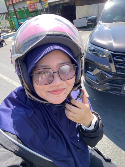 In a selfie taken from the perspective of a motorcycle rider, a woman wearing a purple hijab and glasses is visible. She is wearing a pink and gray helmet with a clear visor. The woman is looking at the camera and smiling slightly. Her left hand is holding the helmet strap, and on her left ring finger, she wears a gold ring with a small stone. A dark-colored car is visible to the right of the motorcycle. In the background, there are buildings, including a shop with a sign that reads "WARUNG MAKAN" (eating place) and another sign with Indonesian text. The scene appears to be on a street in Jawa, Indonesia, indicated by the Indonesian signage. The environment suggests a sunny day.